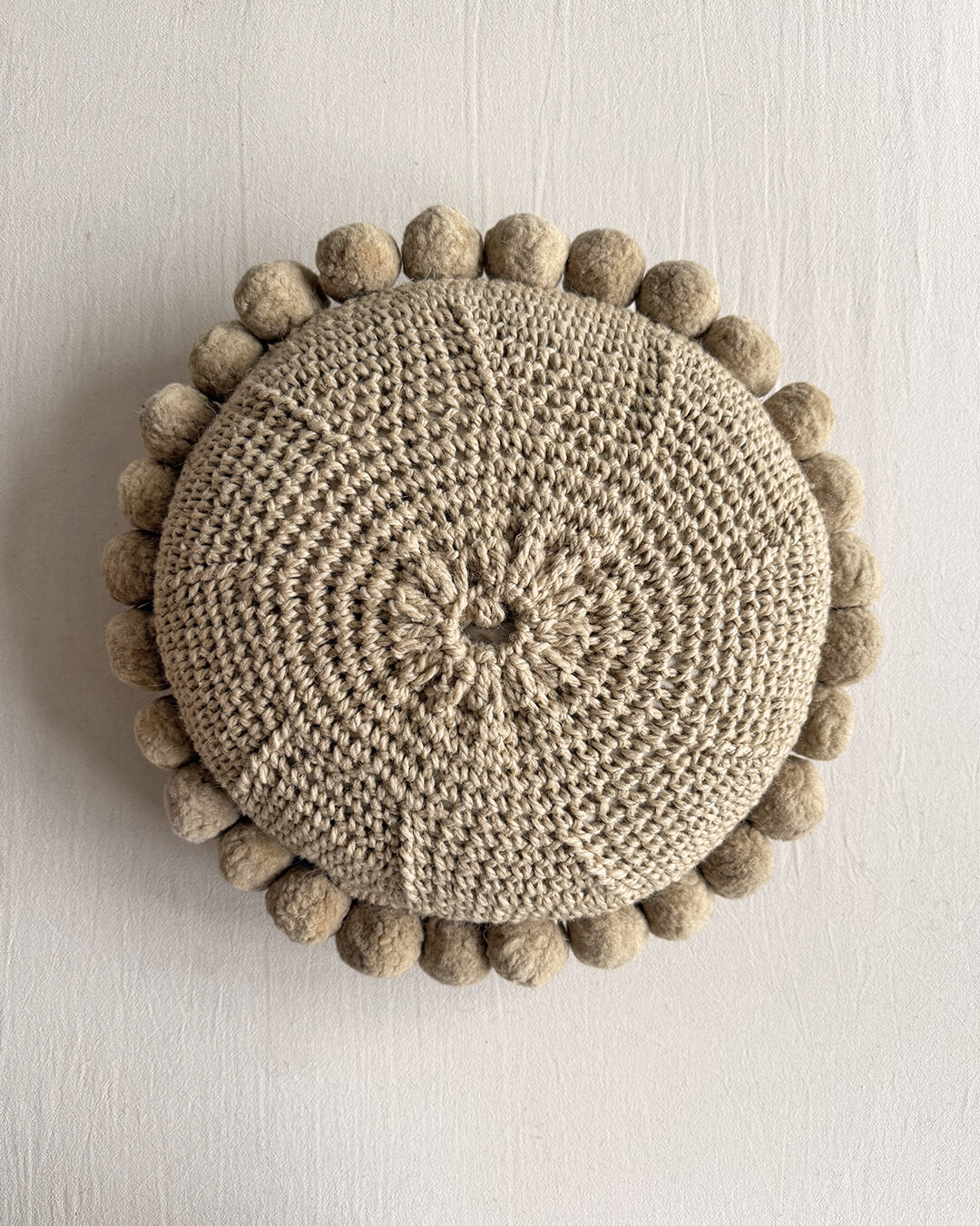 Back view of a Beige-Sand colored round cushion with handmade pom poms.