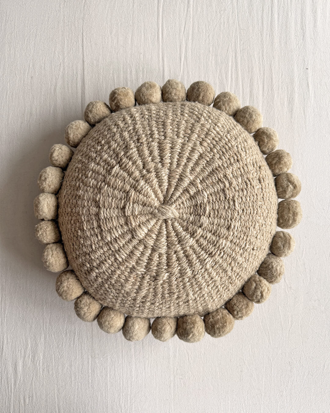 Beige-Sand colored round cushion with handmade pom poms, shown from the front.