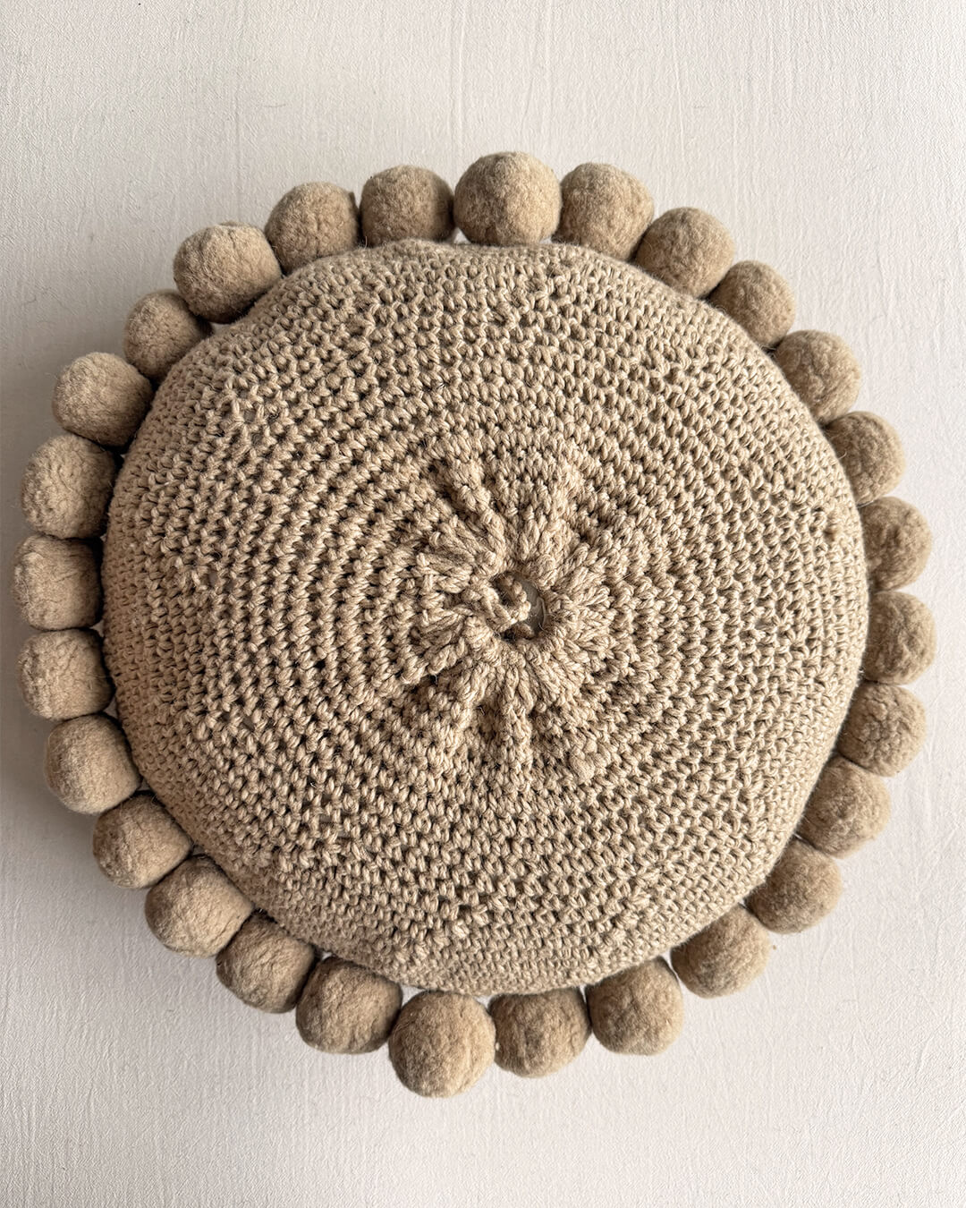 Back view of a Beige-Sand colored round cushion with handmade pom poms.