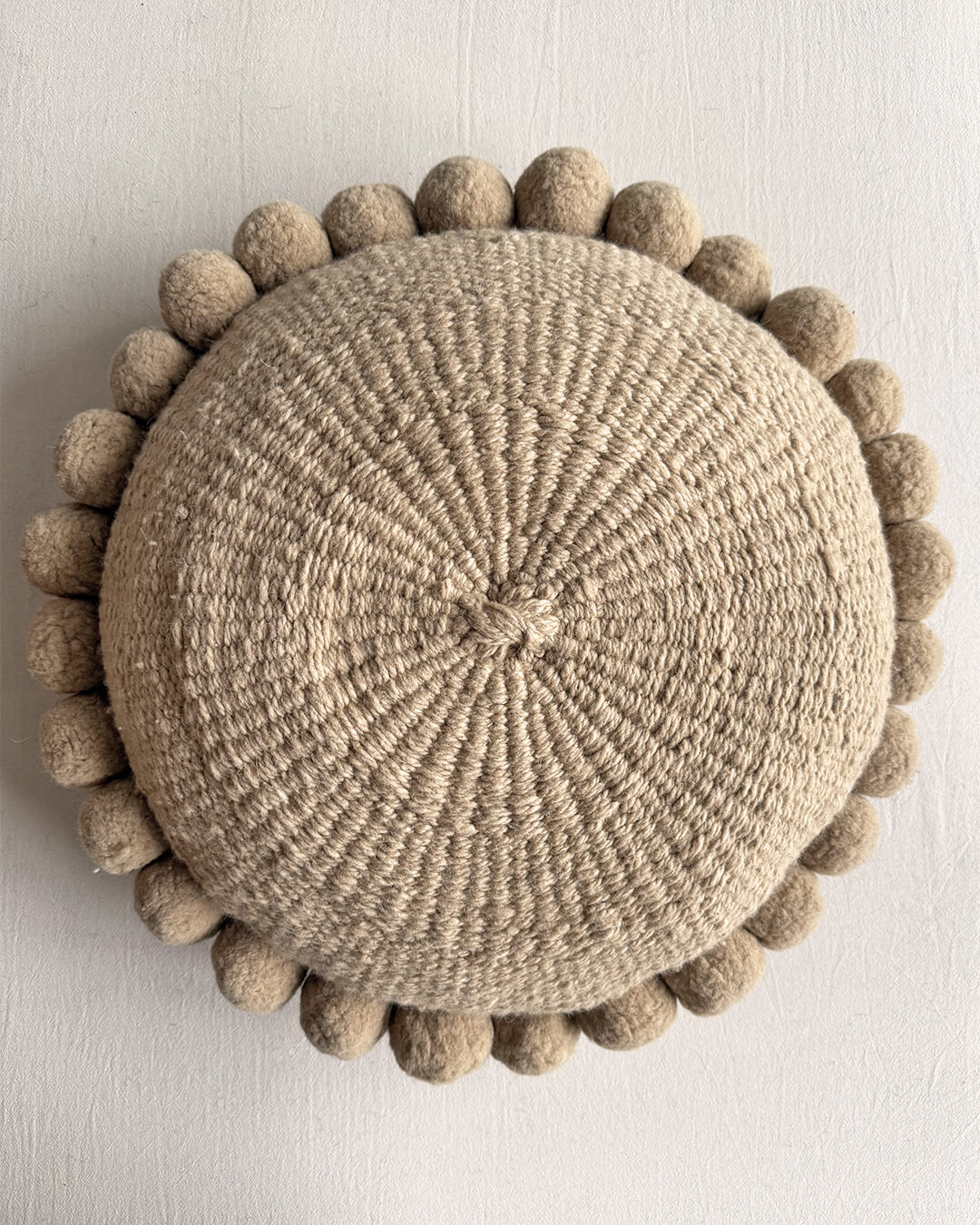 Beige-Sand colored round cushion with handmade pom poms, shown from the front.