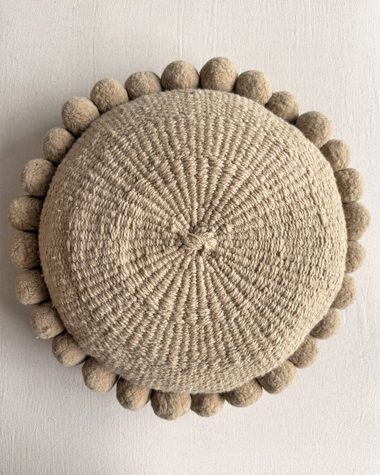Beige-Sand colored round cushion with handmade pom poms, shown from the front.