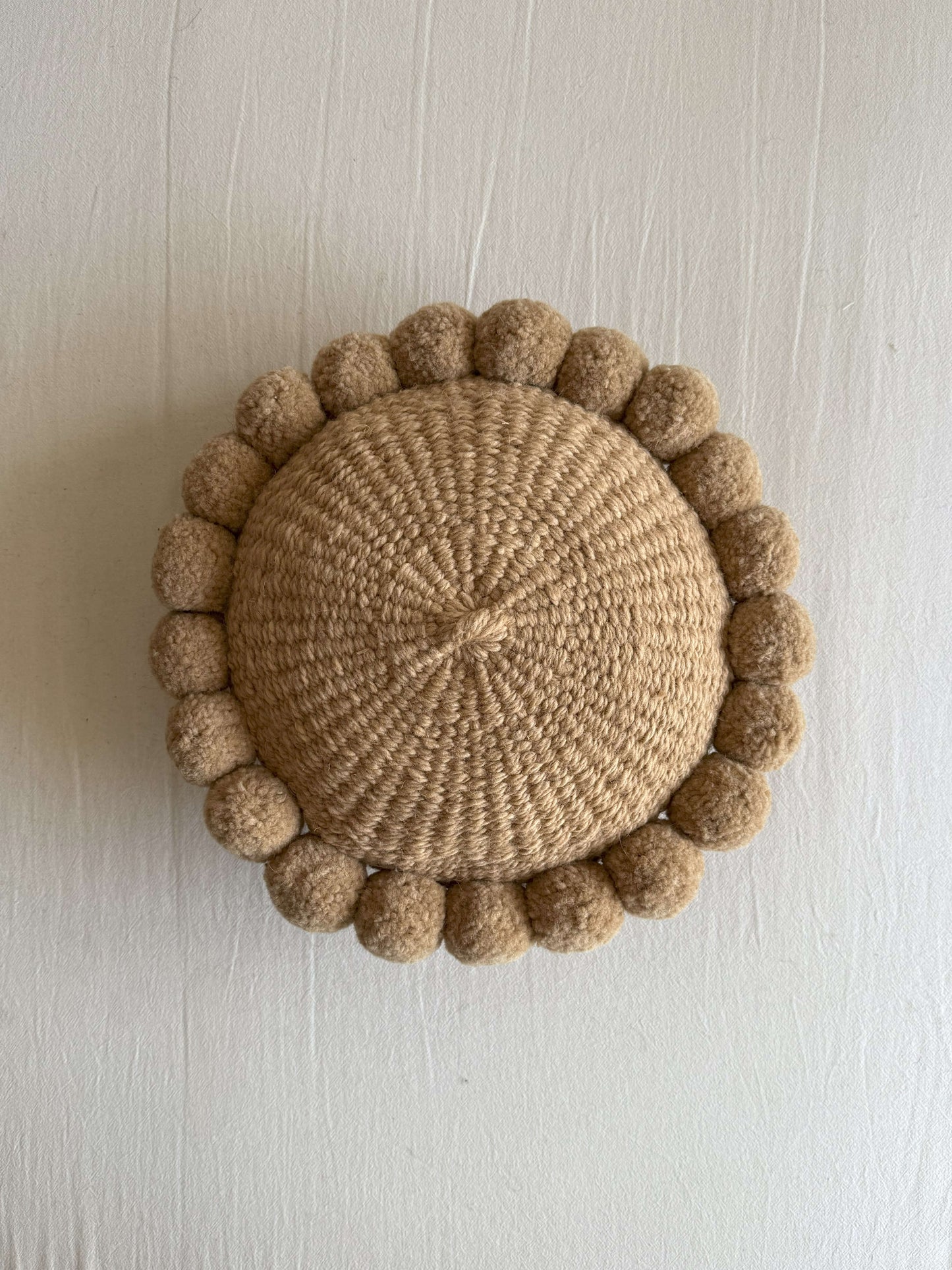 Beige-Sand colored round cushion with handmade pom poms, shown from the front.