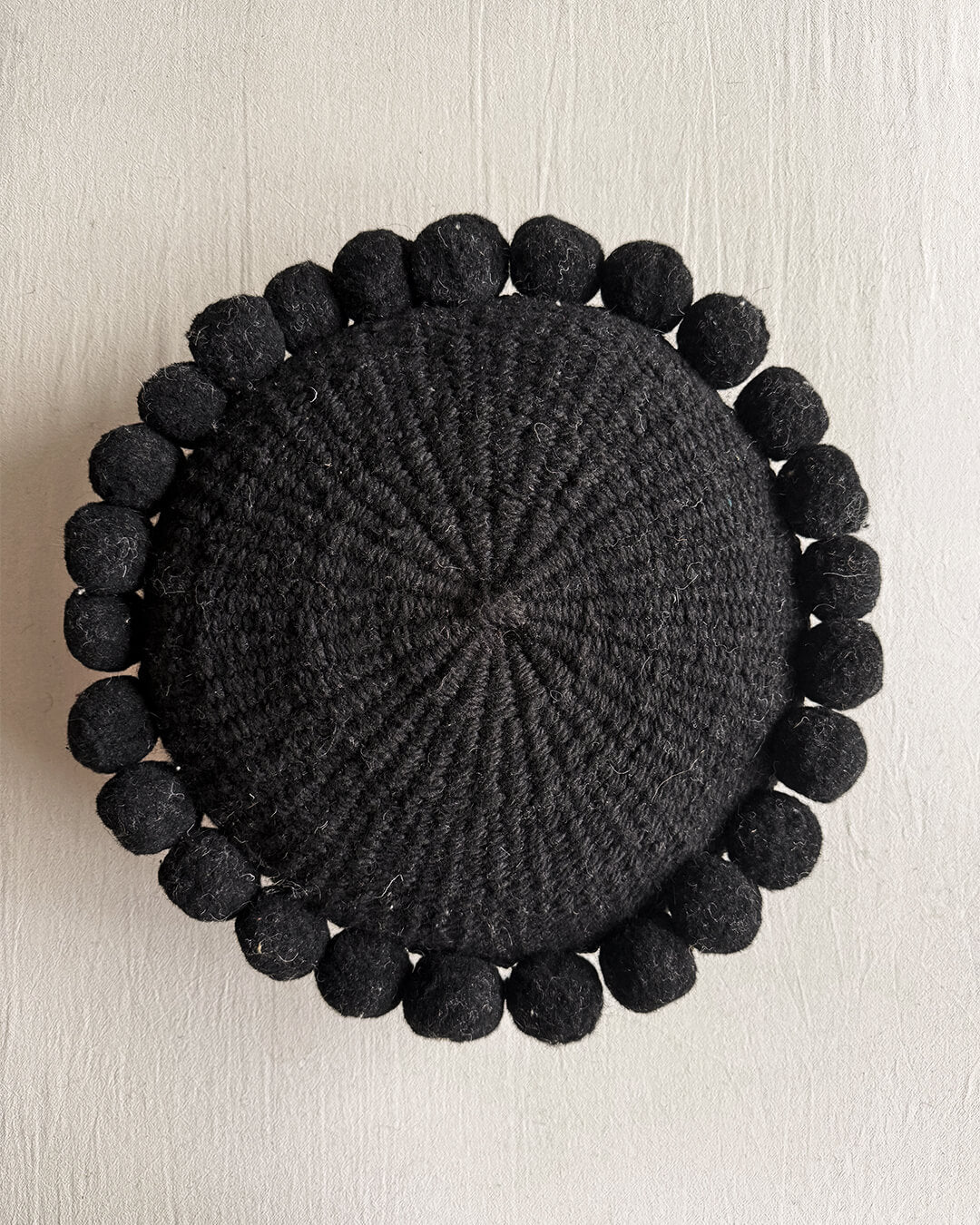 Black-colored round cushion with handmade pom poms, shown from the front.