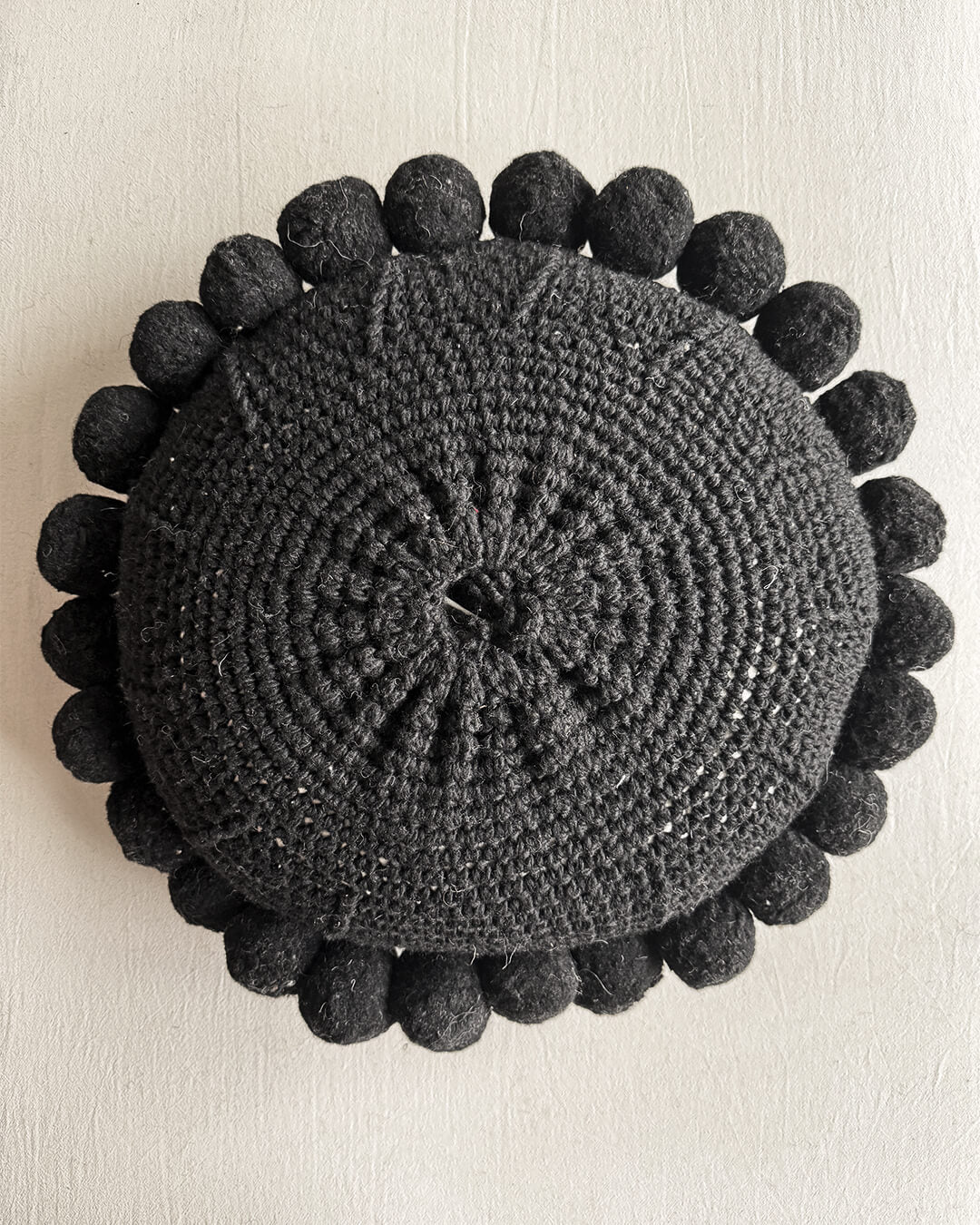 Black-colored round cushion with handmade pom poms, shown from the back.