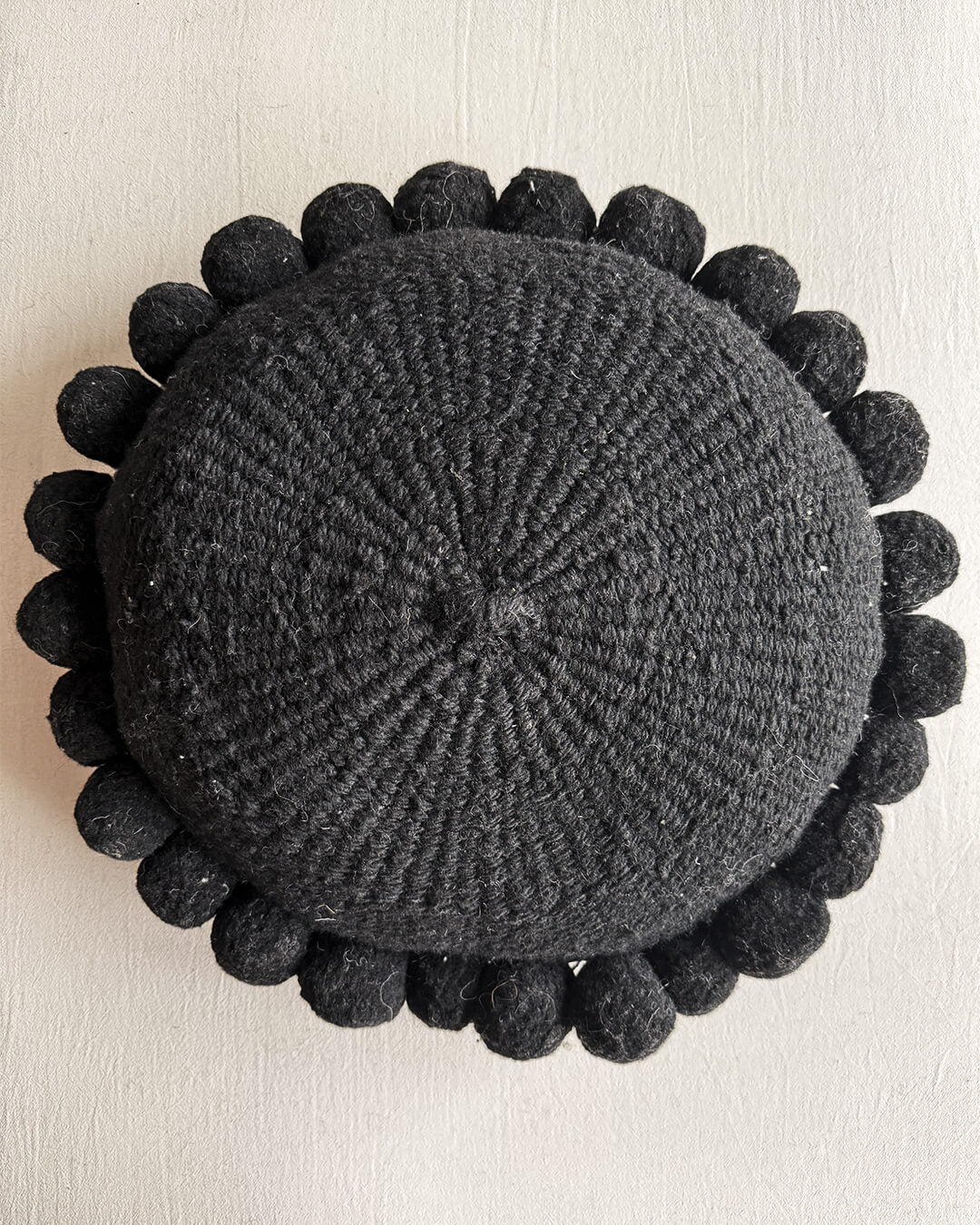 Black-colored round cushion with handmade pom poms, shown from the front.