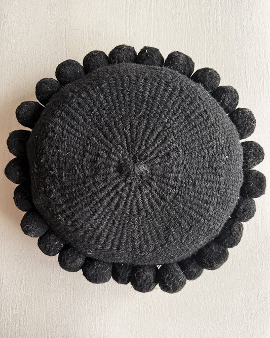 Black-colored round cushion with handmade pom poms, shown from the front.