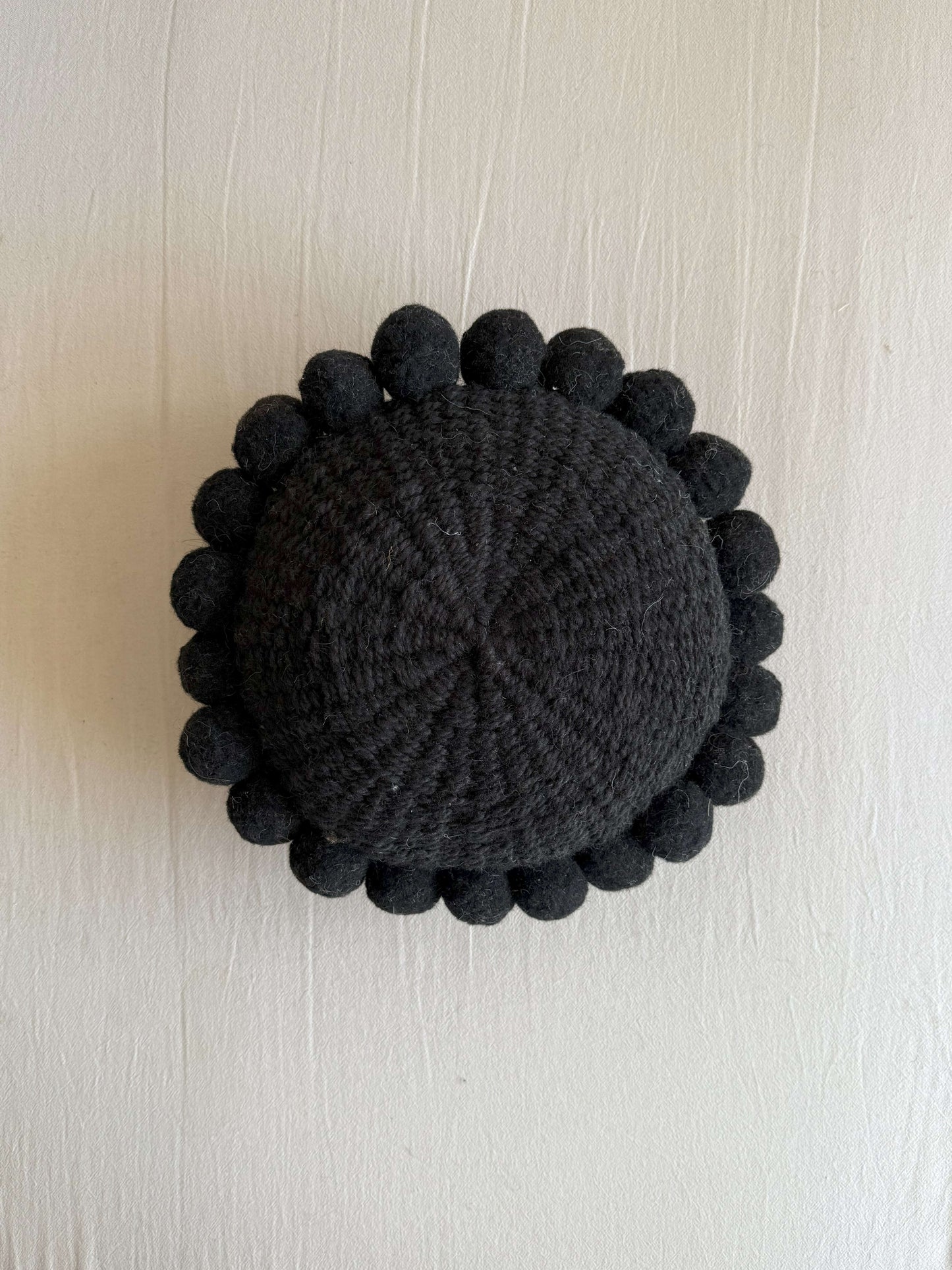 Black-colored round cushion with handmade pom poms, shown from the front.