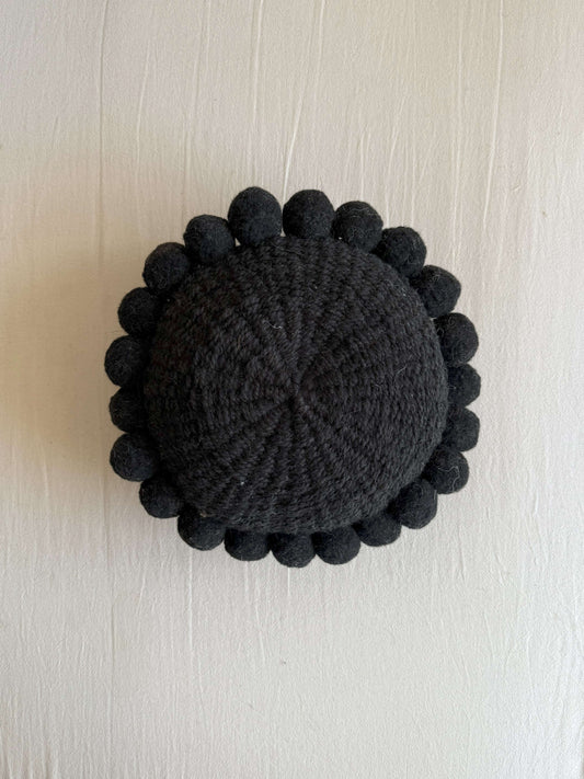 Black-colored round cushion with handmade pom poms, shown from the front.