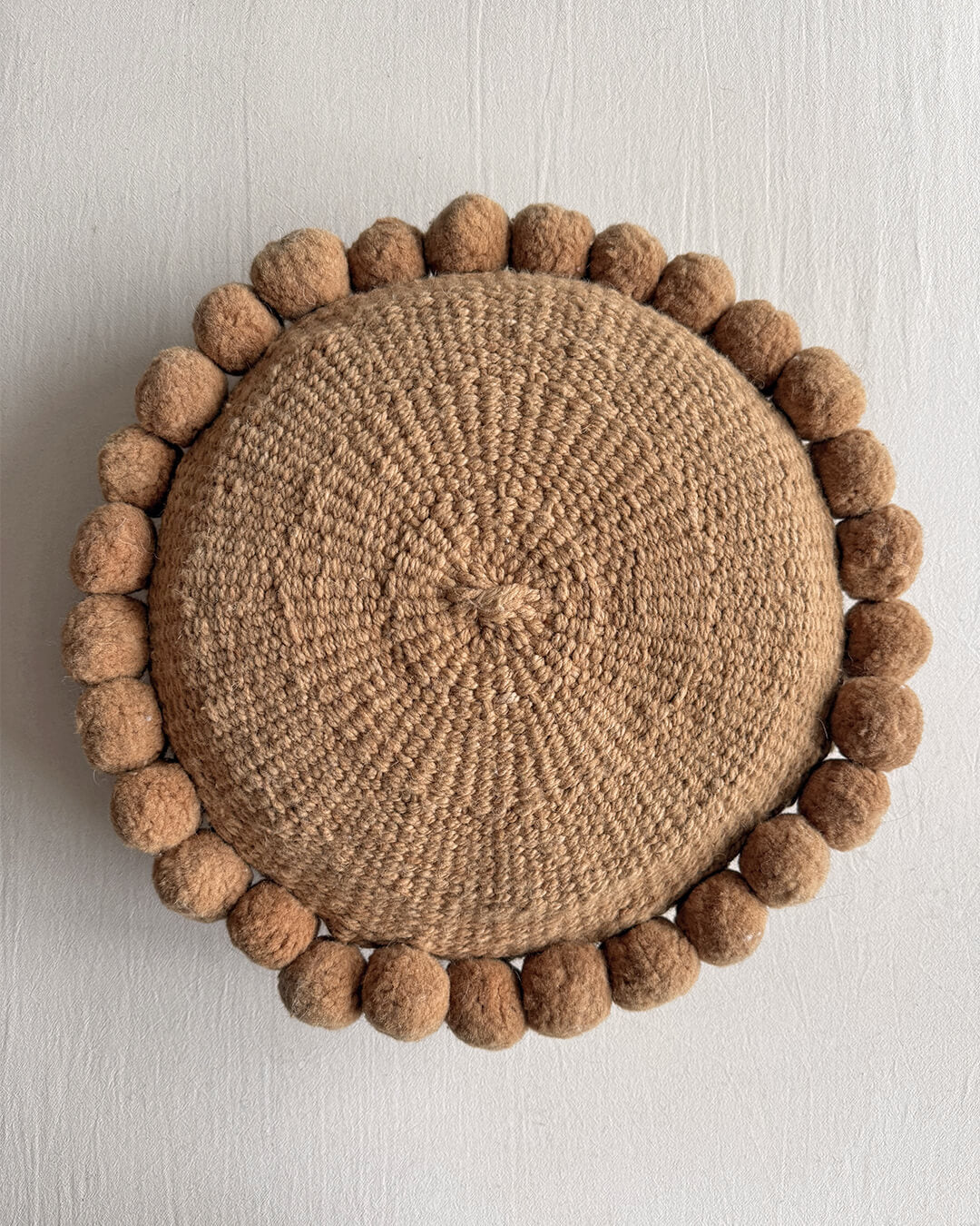 Camel-colored round cushion with handmade pom poms, shown from the front.