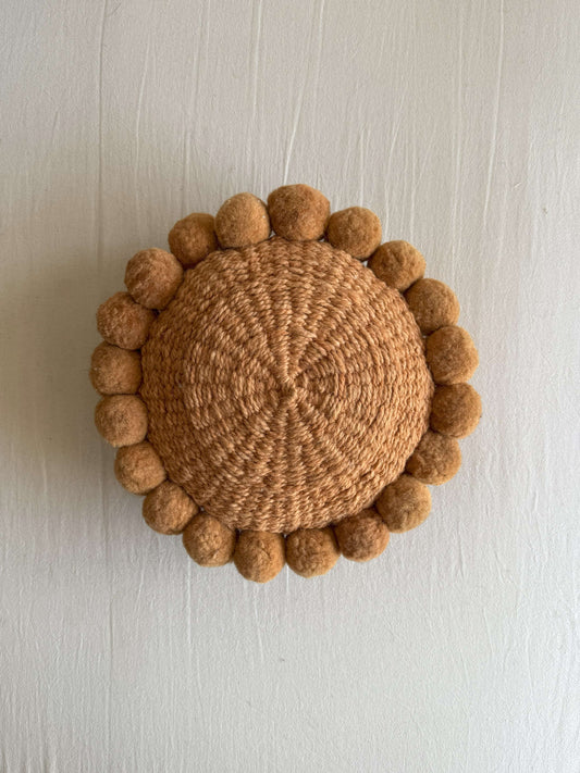 Camel-colored round cushion with handmade pom poms, shown from the front.