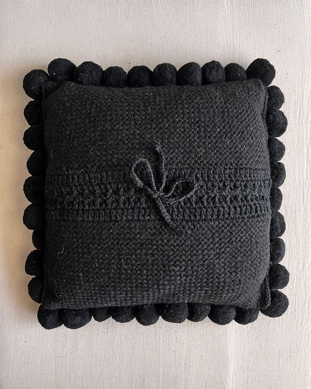 Black-colored square cushion with handmade pom poms, shown from the back.