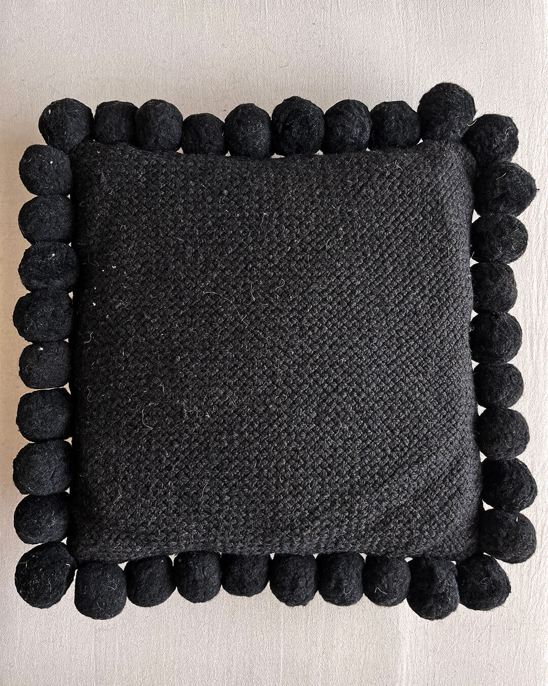 Black-colored square cushion with handmade pom poms, shown from the front.
