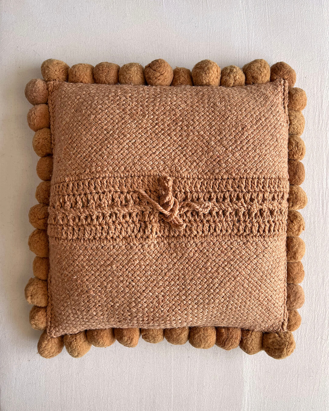 Camel-colored square cushion with handmade pom poms, shown from the back.