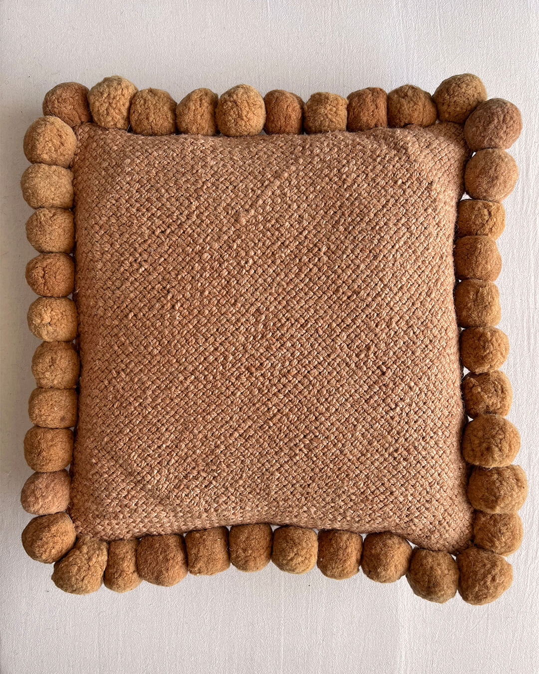 Camel-colored square cushion with handmade pom poms, shown from the front.