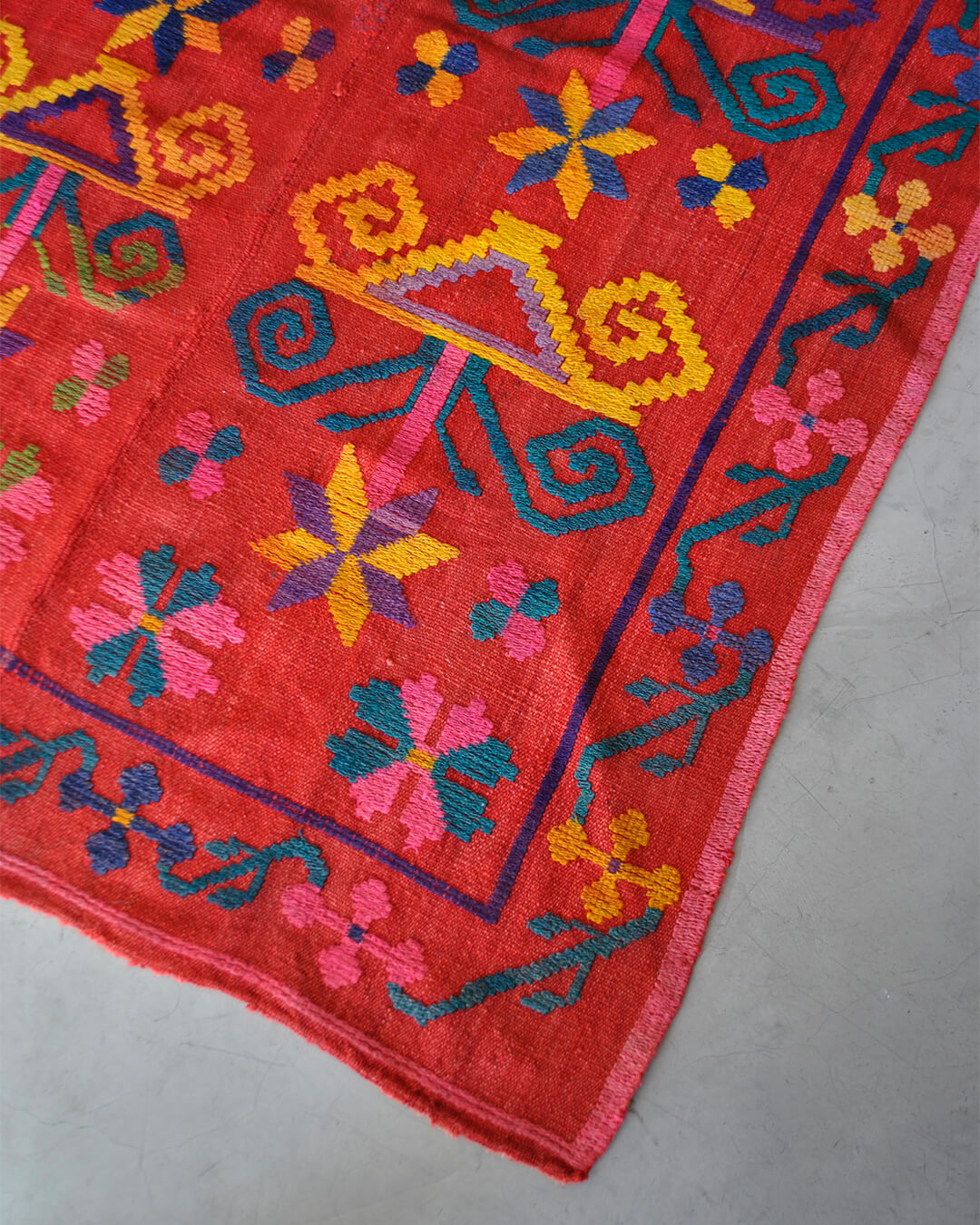 Corner of a red CHACO frazada with colorful handwoven shapes and details in yellow, pink, blue, and green.