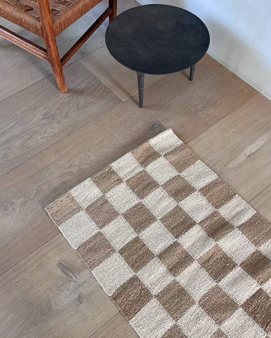Handwoven wool checkered rug in natural and brown colors placed on a wooden floor with a chair and a round stool nearby.