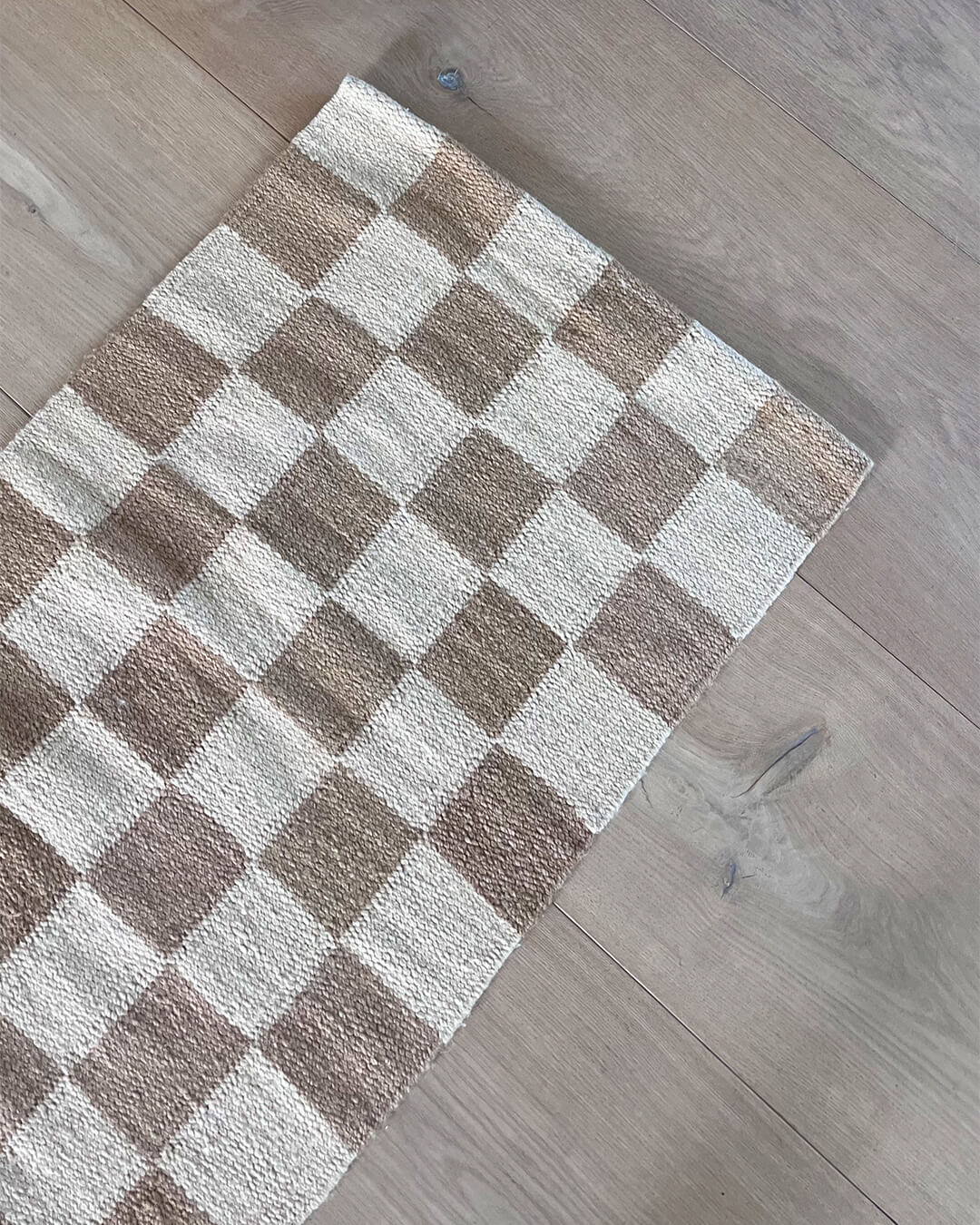 Top view of a handwoven wool runner rug with a checkered pattern in natural and brown tones on a wooden floor.