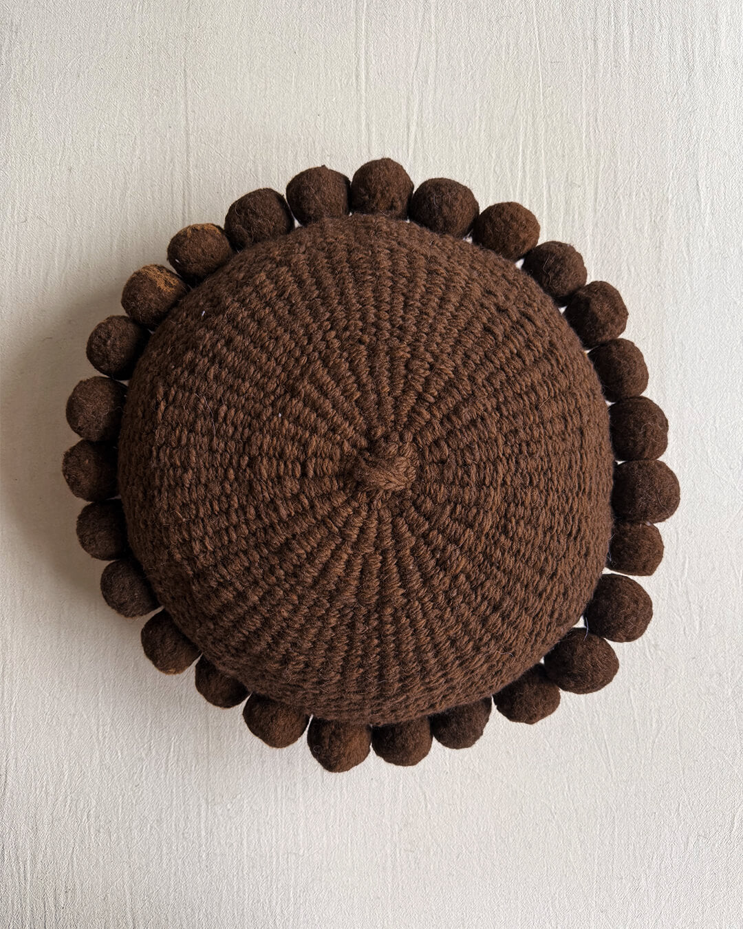 Chocolate Brown-colored round cushion with handmade pom poms, shown from the front.