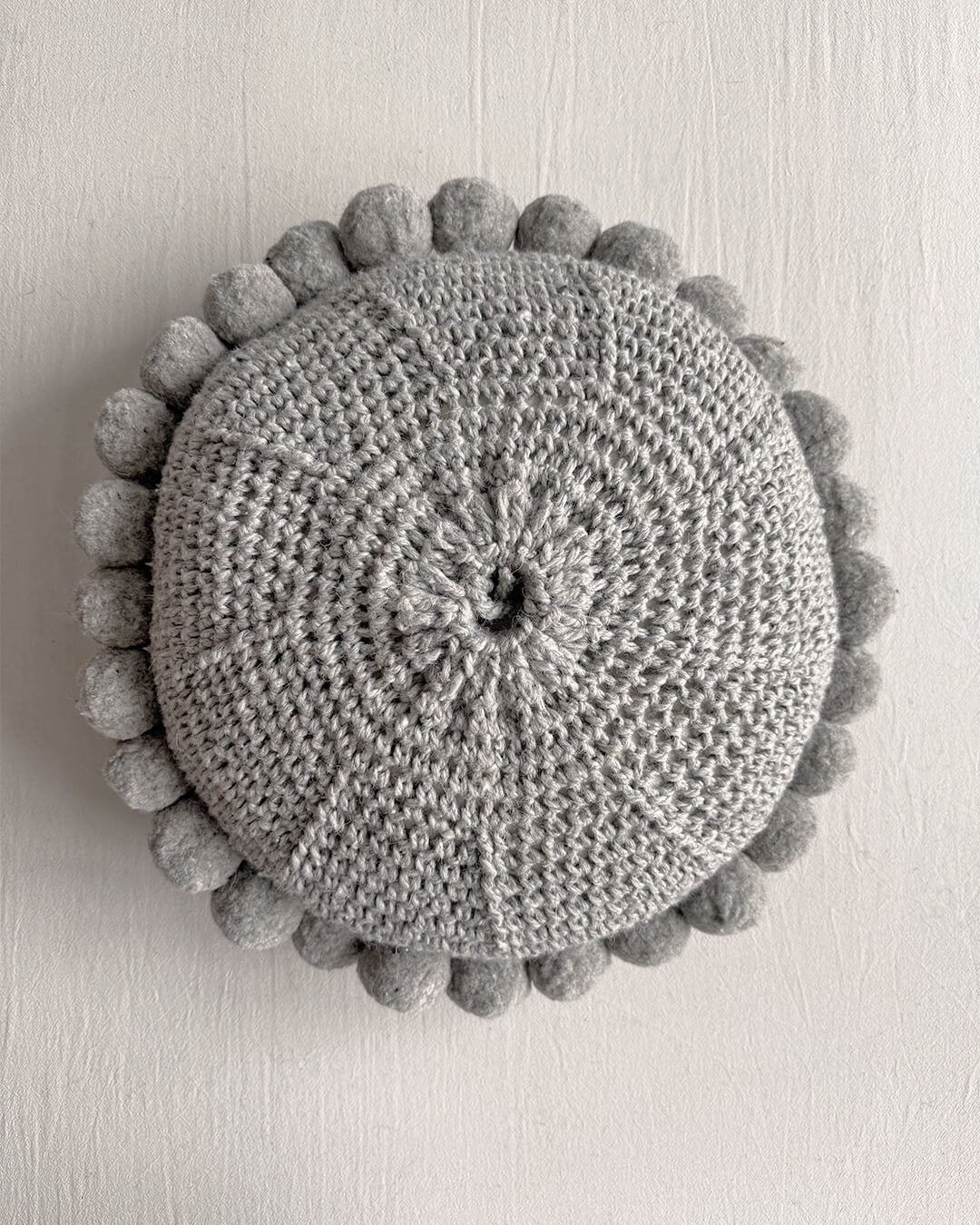 Gray-colored round cushion with handmade pom poms, shown from the back.