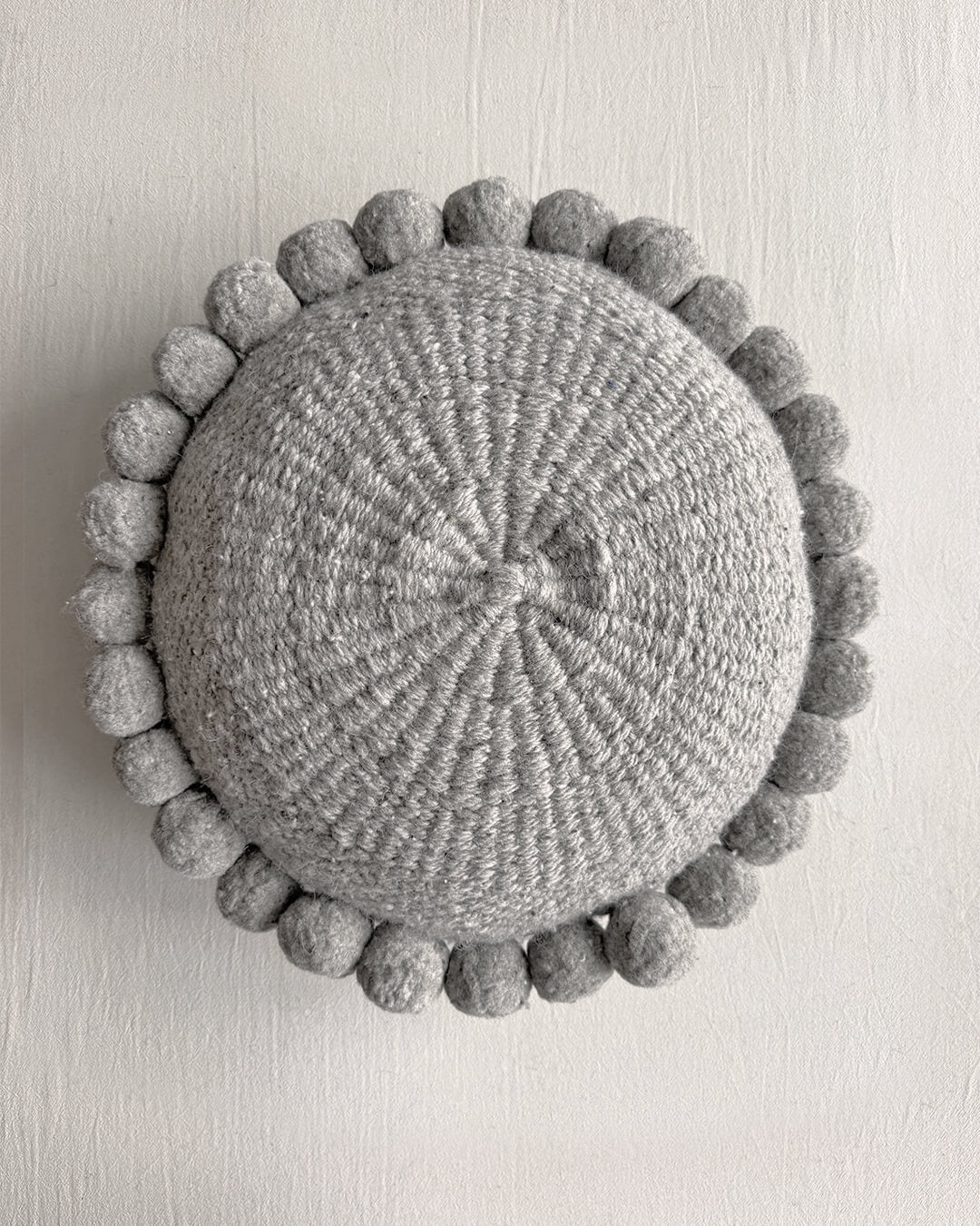 Gray-colored round cushion with handmade pom poms, shown from the front.