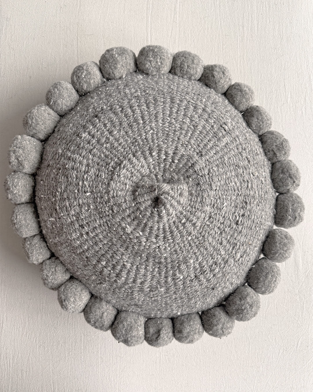 Gray-colored round cushion with handmade pom poms, shown from the front.