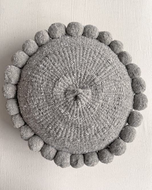 Gray-colored round cushion with handmade pom poms, shown from the front.