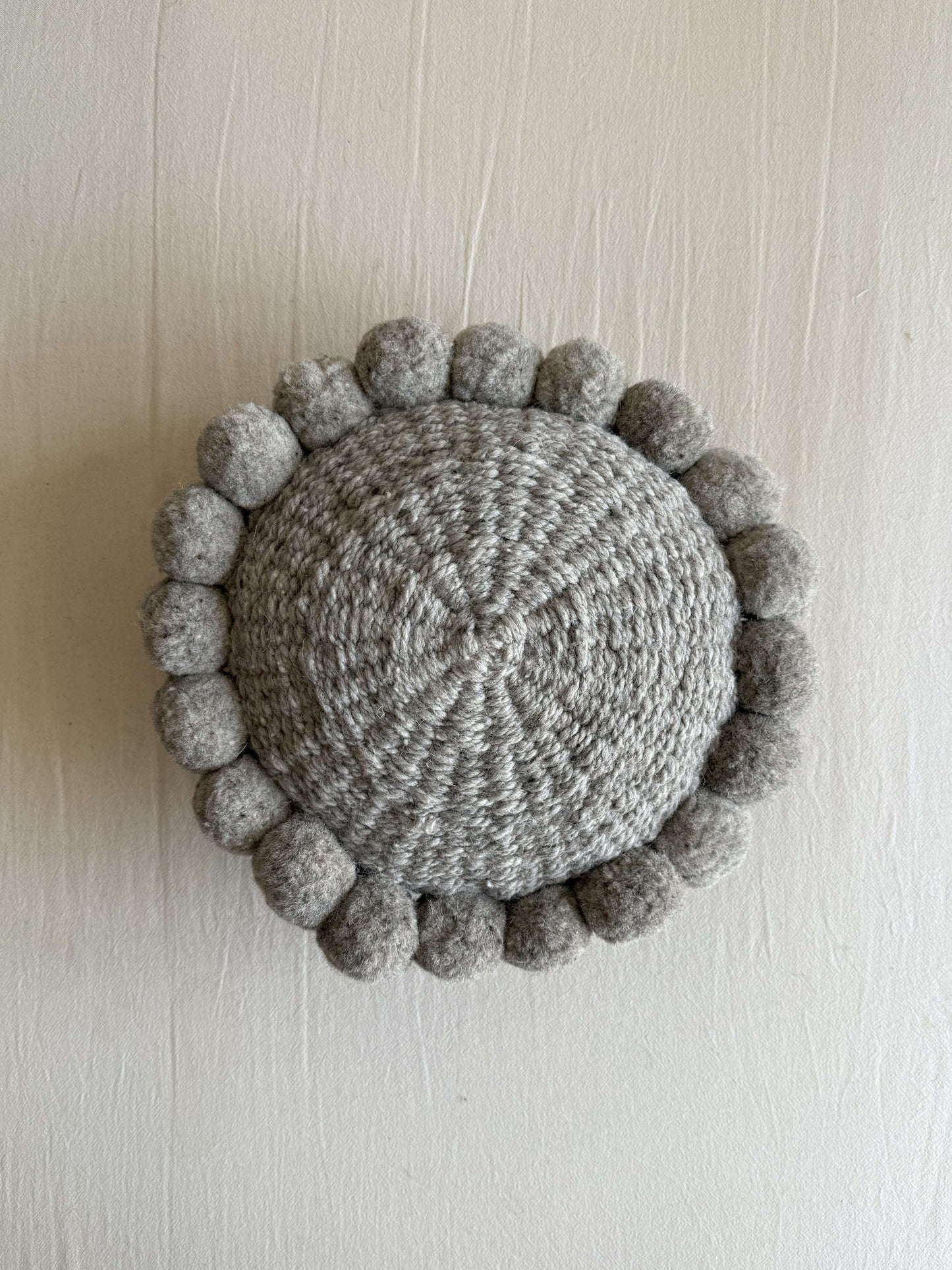 Gray-colored round cushion with handmade pom poms, shown from the front.