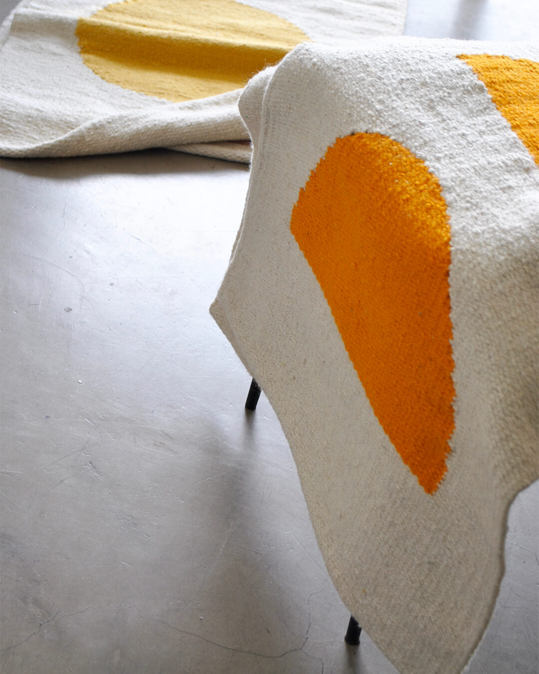 Handwoven CHACO Mini Rugs with sun-inspired designs in yellow and orange tones, displayed on a light concrete floor.