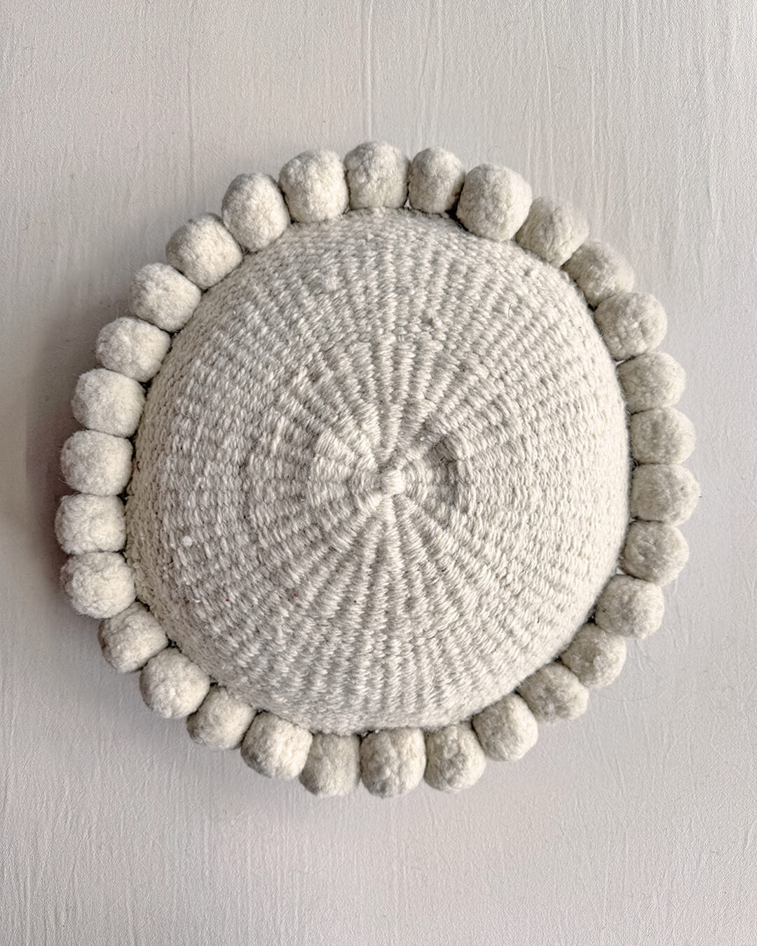 Natural White-colored round cushion with handmade pom poms, shown from the front.