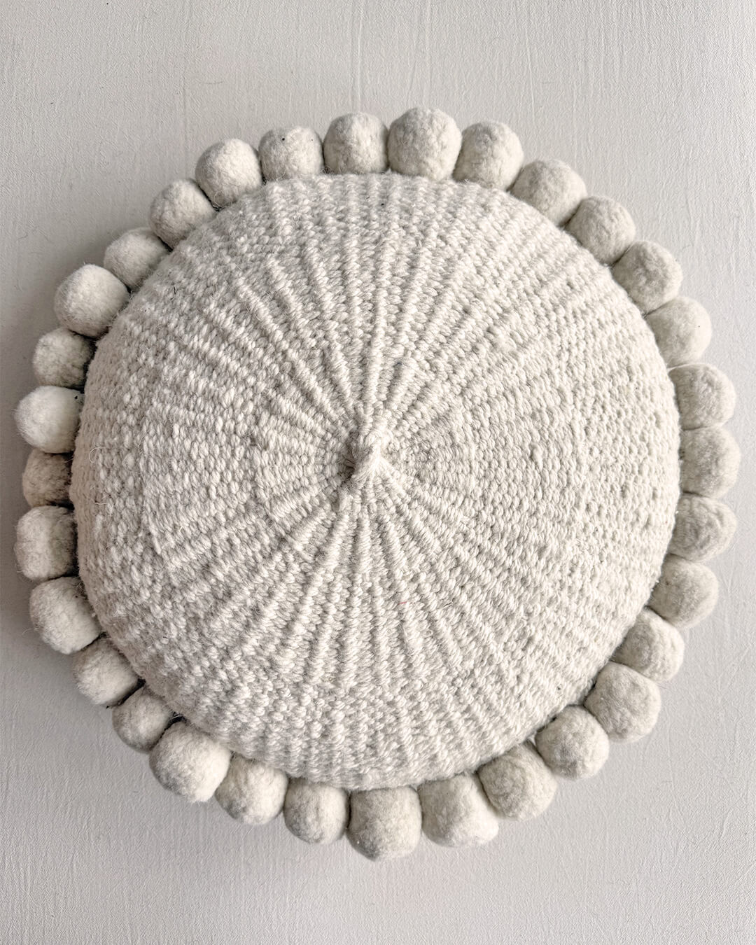 Natural White-colored round cushion with handmade pom poms, shown from the front.