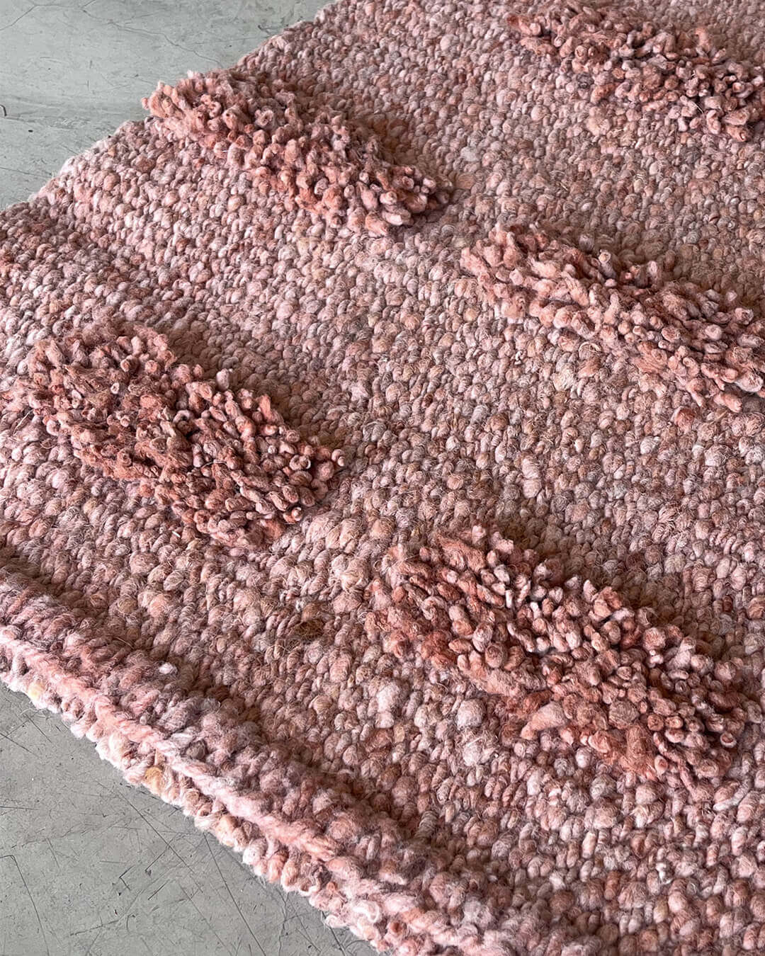 Close-up of a handwoven wool runner rug in old rose color featuring raised pom-pom texture on a light concrete floor.