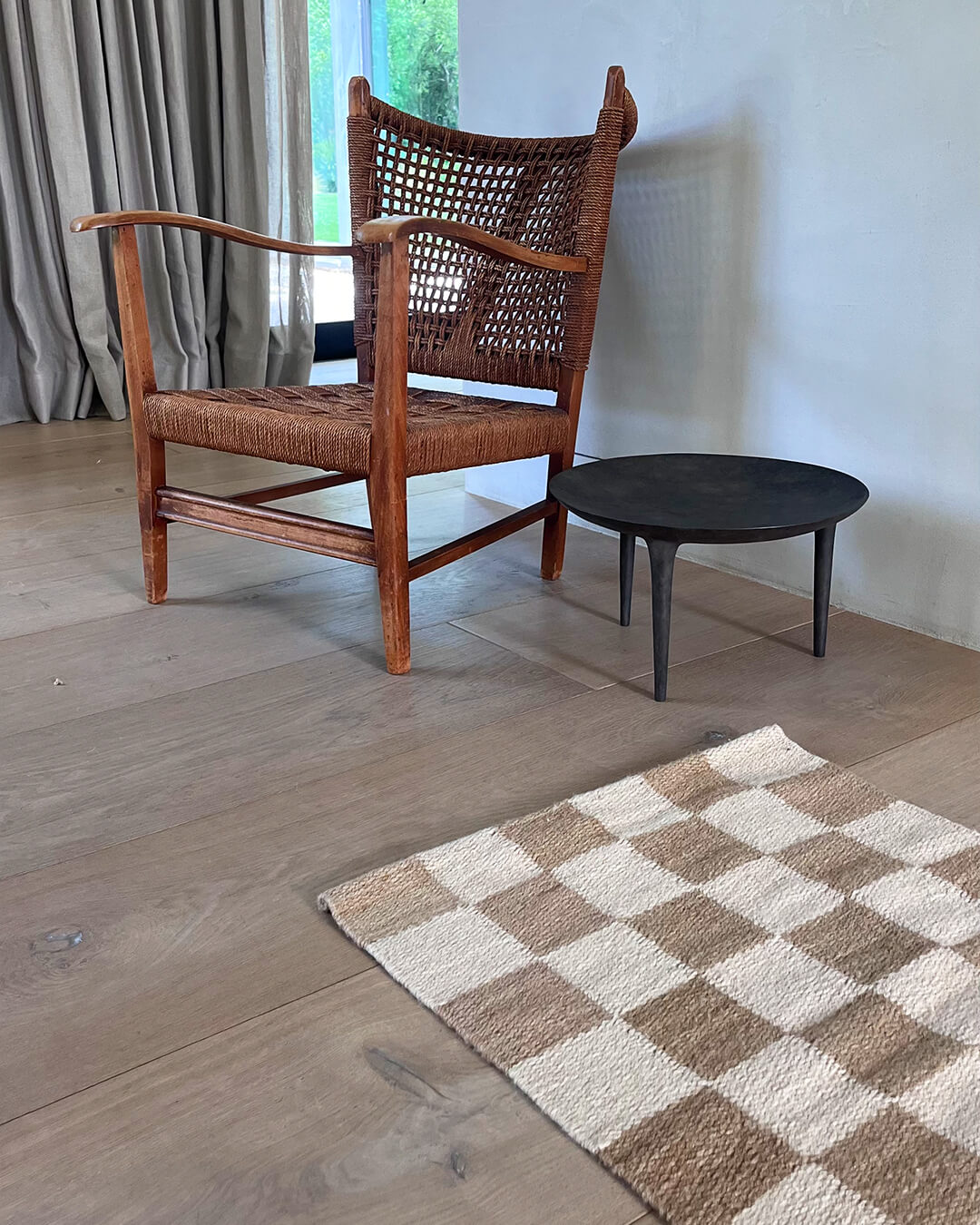 Handwoven wool checkered rug in natural and brown tones styled on a wooden floor beside a woven wooden armchair and a small round side table.