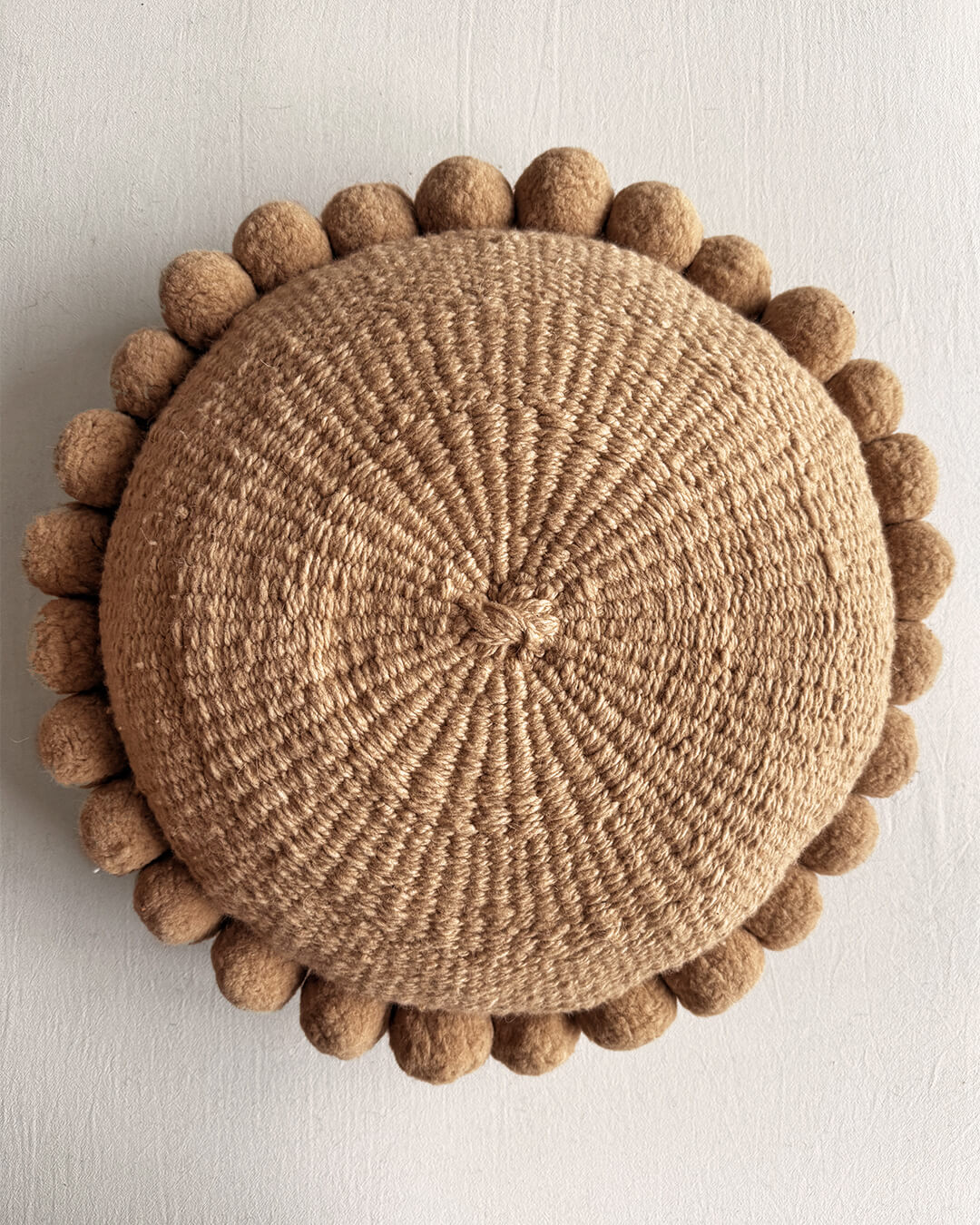 Camel-colored round cushion with handmade pom poms, shown from the front.
