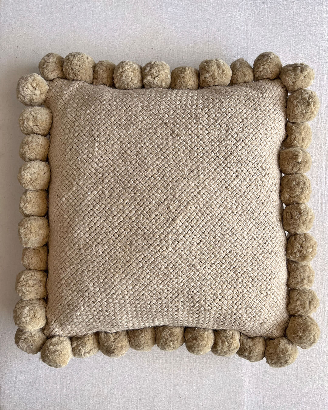 Beige-Sand colored square cushion with handmade pom poms, shown from the front.