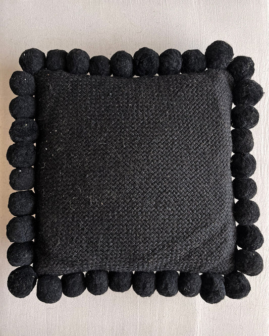 Black-colored square cushion with handmade pom poms, shown from the front.