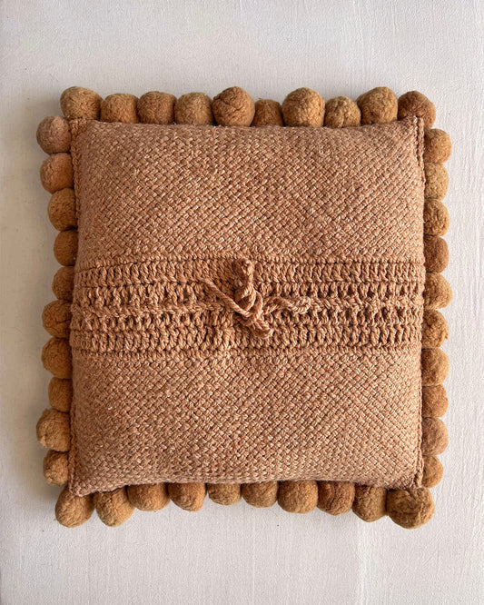 Camel-colored square cushion with handmade pom poms, shown from the back.