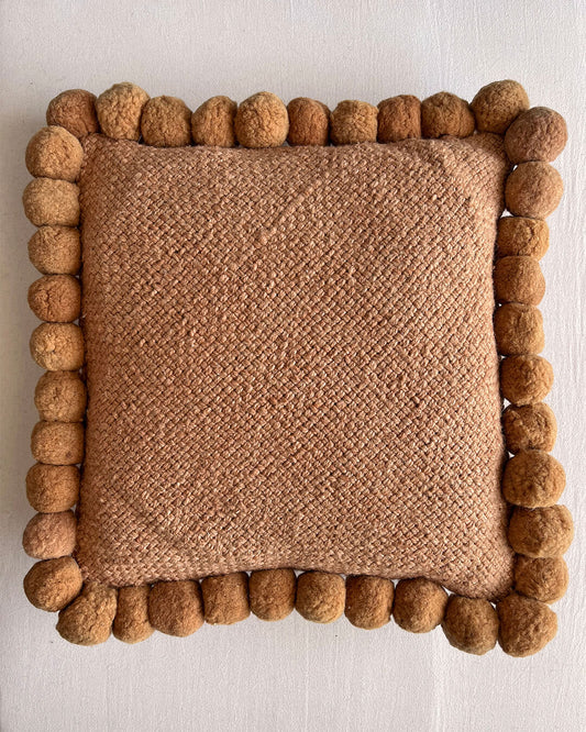 Camel-colored square cushion with handmade pom poms, shown from the front.