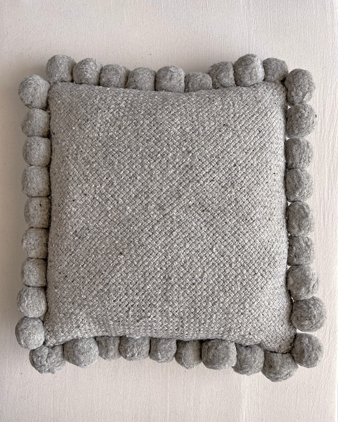 Gray-colored square cushion with handmade pom poms, shown from the front.