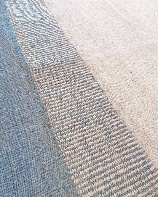 Close-up of a handwoven wool rug showing blue-gray and natural beige sections with a textured woven border detail.