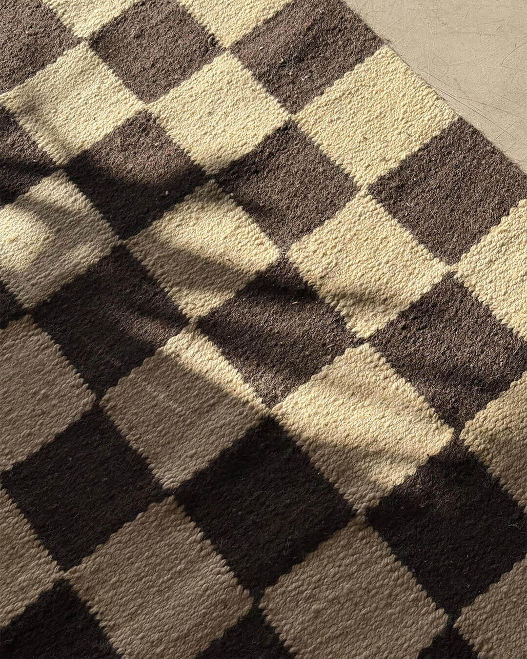 Close-up of a handwoven wool rug with a checkered pattern in black and sand tones, showing the textured surface under natural light.