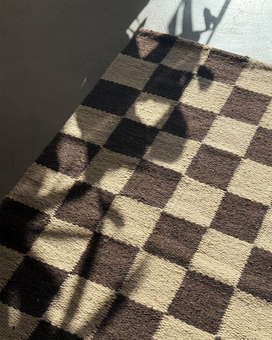 Handwoven wool rug with a black and sand checkered design placed on the floor, partially lit by soft natural shadows.