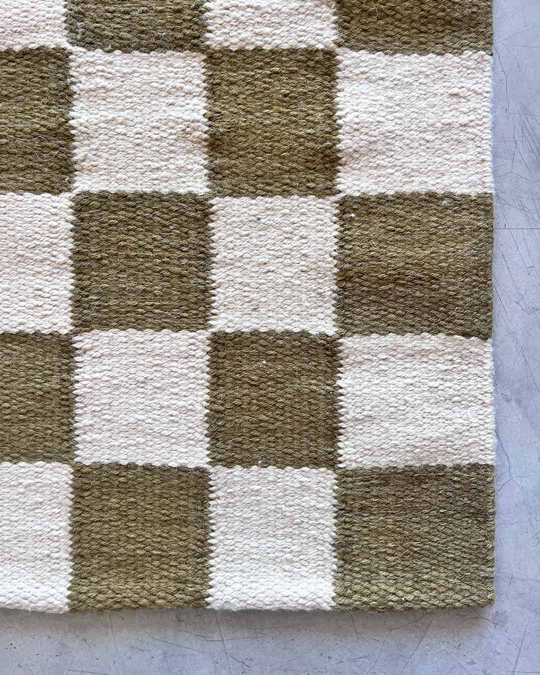 Top view of a handwoven wool rug with sand and moss green checkered squares placed on a concrete floor.