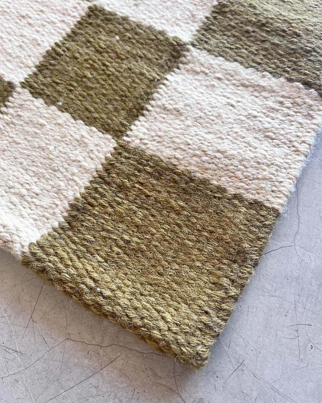 Detail view of the corner of a handwoven wool rug showing a sand and moss checkered pattern and thick woven edge.