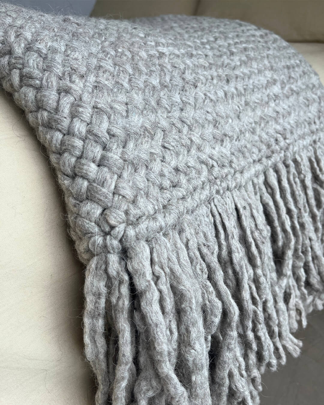 Gray wool throw draped over the side of a light sofa, showing weave and fringe.