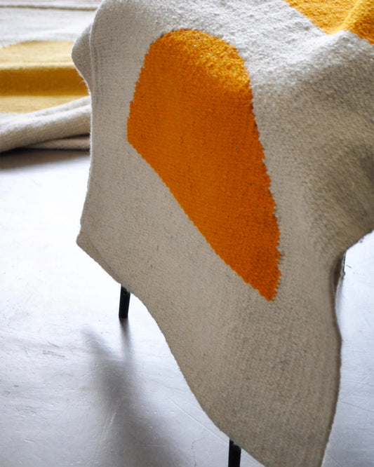 Handwoven CHACO Mini Rug draped over a chair, featuring an orange sun design on a white wool base.
