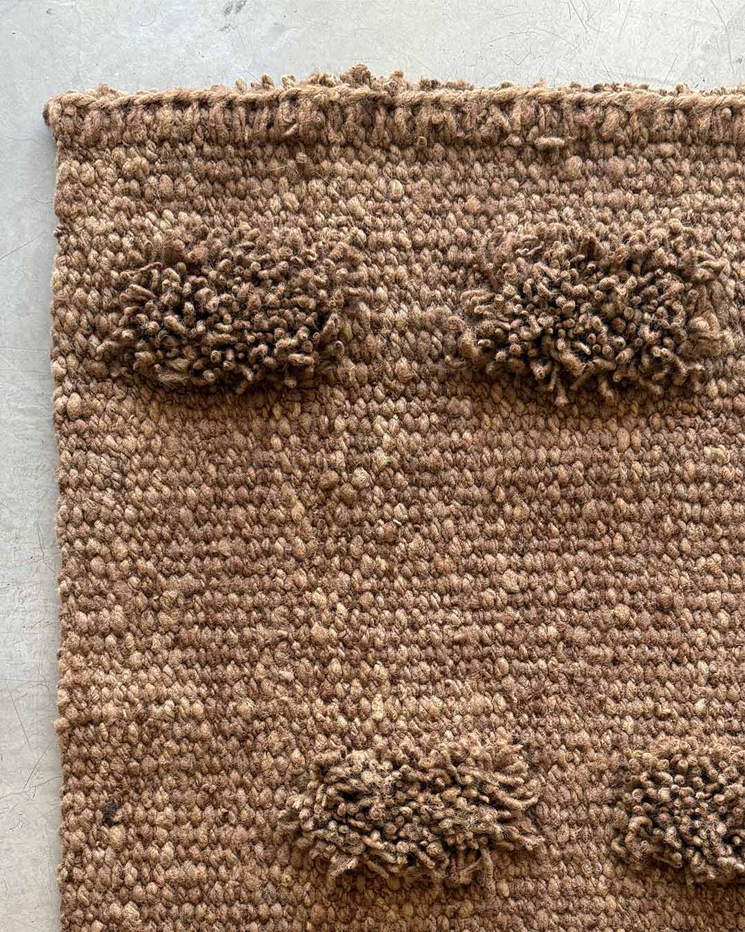 Close-up of a handwoven wool runner rug in wood brown with raised pom-pom details against a concrete background.