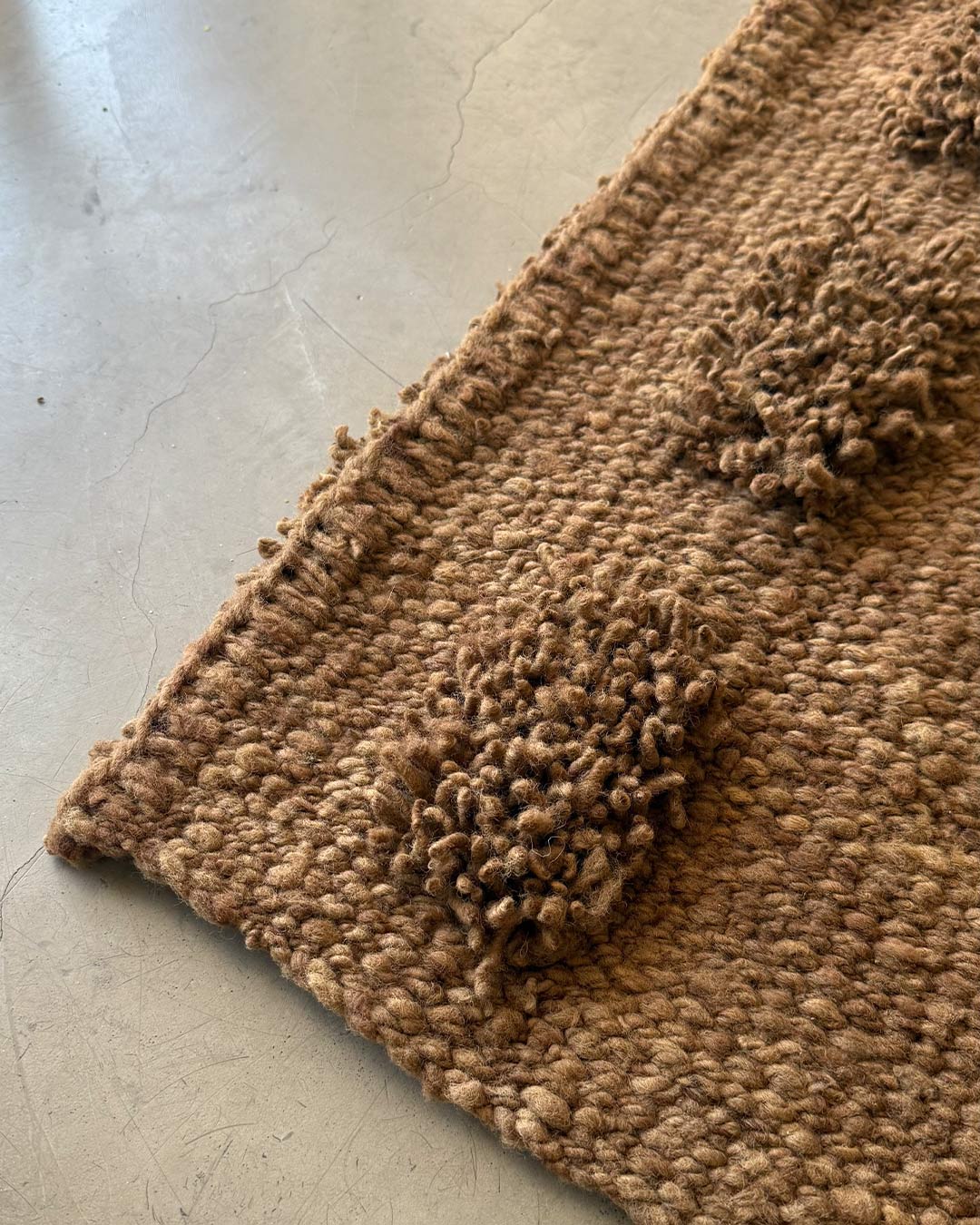 Corner detail of a handwoven wool runner rug in wood brown, showing the pom-pom texture and woven edge on a concrete floor.