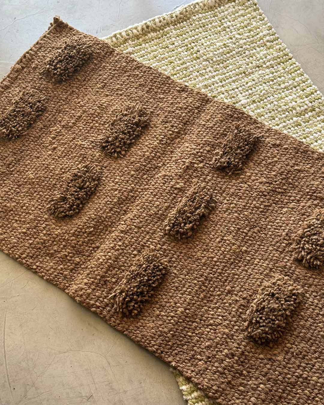 Handwoven wool runner rug in wood brown with raised pom-pom texture, layered over another textile on a light concrete floor.