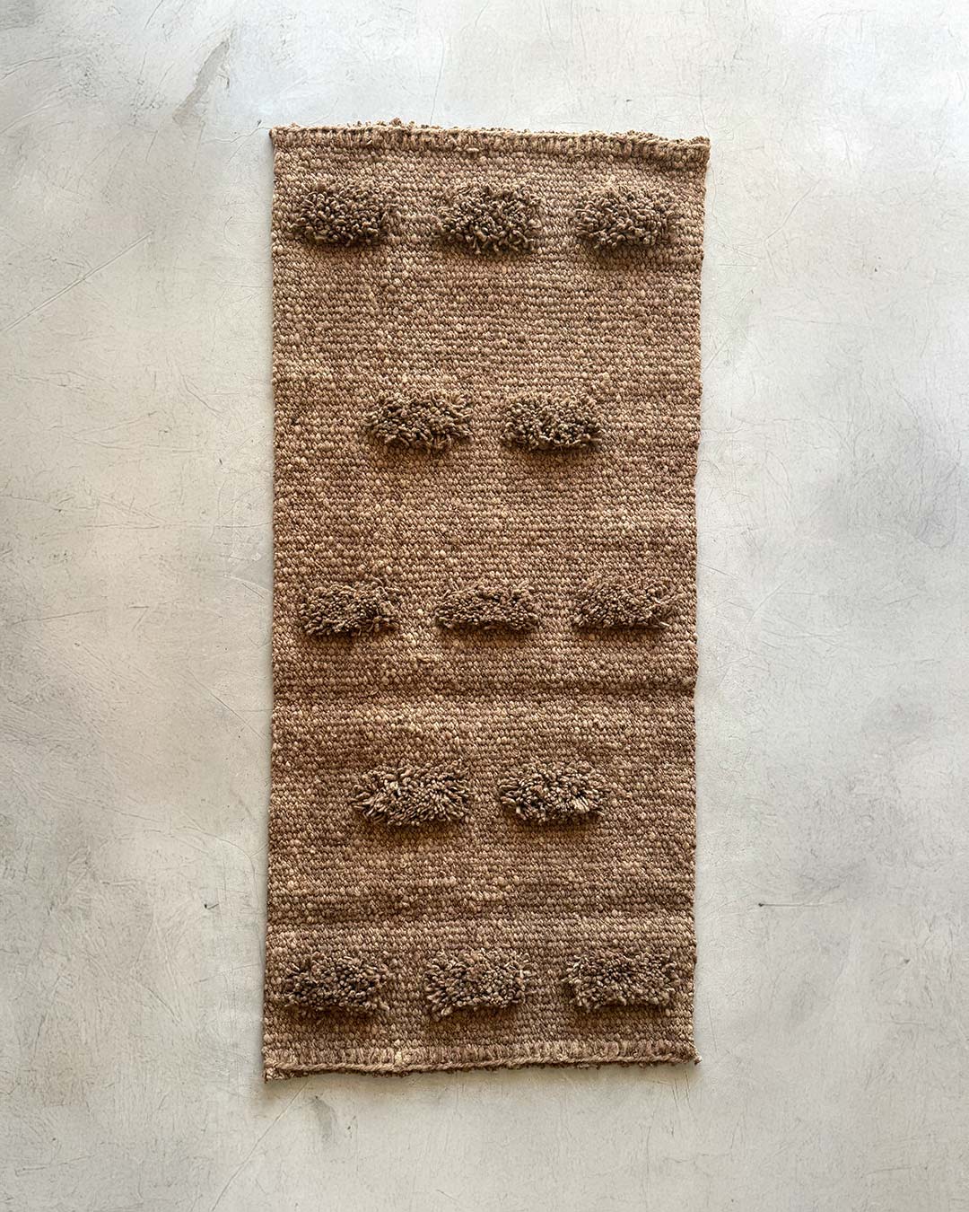 Full view of a handwoven wool runner rug in wood brown with evenly spaced pom-pom texture, photographed on a light concrete surface.