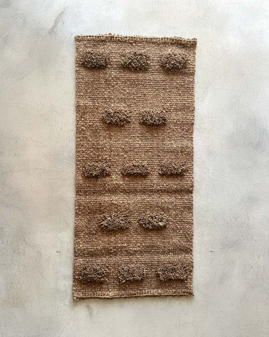 Full view of a handwoven wool runner rug in wood brown with evenly spaced pom-pom texture, photographed on a light concrete surface.